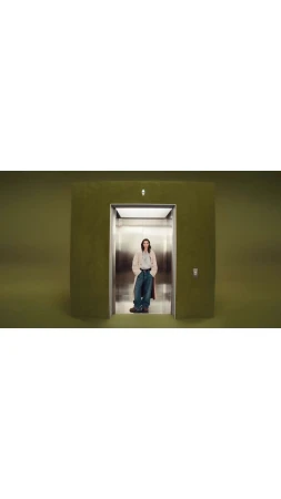 An adult stands in an elevator with an olive green exterior, wearing a pale jacket and jeans, against a solid olive green backdrop.