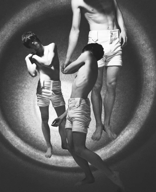 Monochrome art photography of three slender, shirtless adult males with blurred faces wearing shorts against a grainy, circular backdrop.