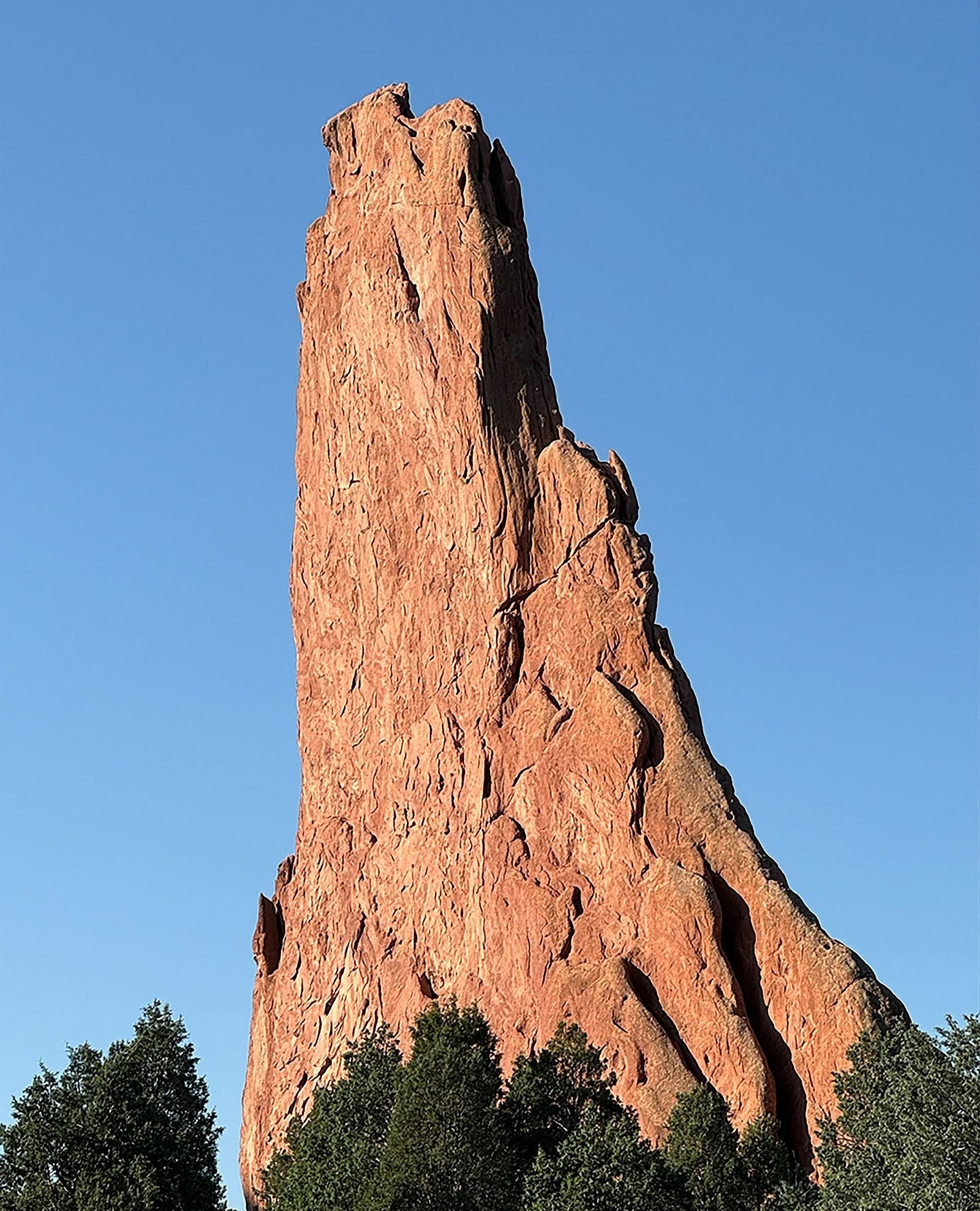 Towering red rock formation against a clear blue sky with treetops visible at the base of the rock, highlighting natural beauty.