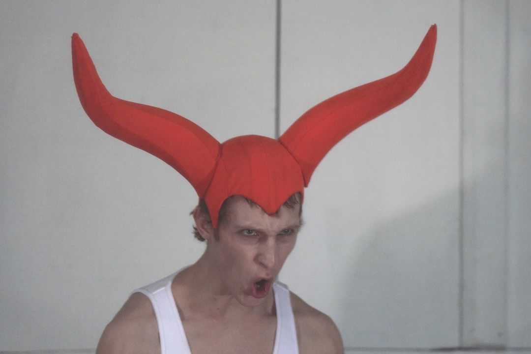 Close-up of a fair-skinned adult male with an open mouth wearing a red horned headdress against a white wall in an indoor setting.