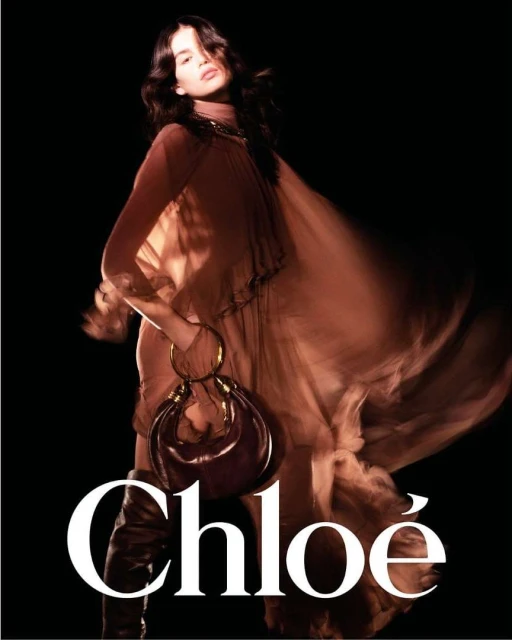 Fashion shot of a woman with long dark hair, wearing a flowing caramel colored dress and boots, holding a rounded handbag, on black backdrop.