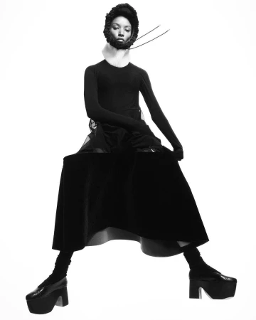 Model in black long sleeve top and long skirt wearing platform shoes and dark bonnet against white background