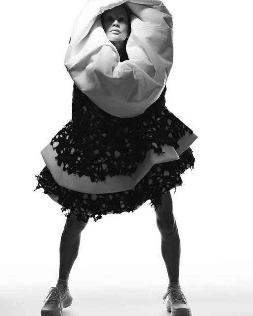 Monochrome fashion portrait of an adult woman wearing an abstract gown with a large hoodie and platform sneakers against a white backdrop.