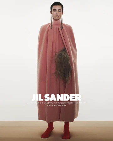 Model in a long pink cape coat with dangling earrings and red boots standing against a neutral studio background