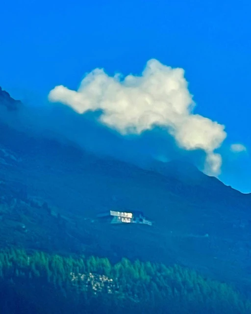 Scenic view of a modern building nestled on a forested mountain with a fluffy cloud in the blue sky above on a sunny day.