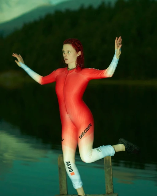 Red-haired woman in red and white wetsuit poses on wooden steps near a lake surrounded by trees and mountains in the background.