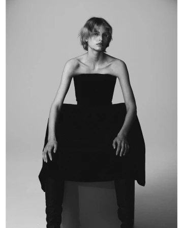 Model with short tousled hair wearing a strapless black dress seated against a plain grey studio backdrop