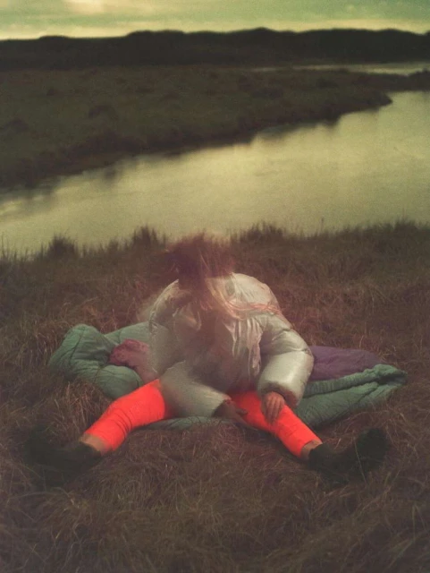 A person in a sleeping bag sits on a grassy hillside near a river under an overcast sky, creating a serene outdoor camping scene.