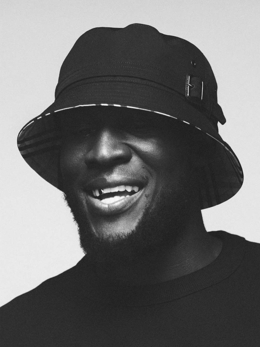 Black and white portrait of a smiling black man wearing a bucket hat and dark t-shirt against a light studio background