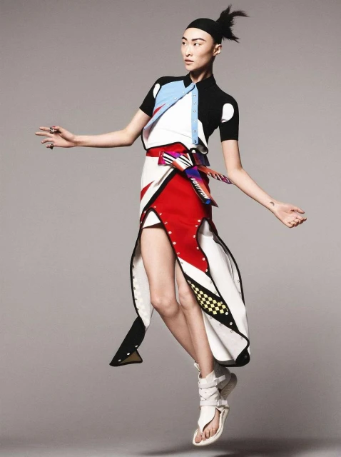 Fashionable Asian woman in colorful geometric outfit strikes a dynamic pose against a grey backdrop in a full-length studio shot.
