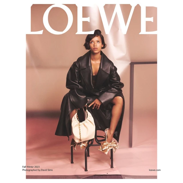 Fashion editorial featuring a model in a black coat, cream-colored bag with chain, and heeled shoes with bows, set against a pale pink backdrop.