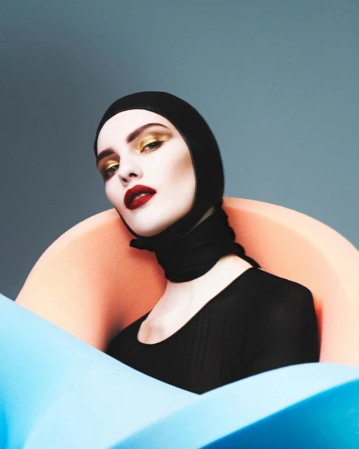 Fashionable woman in black headscarf with bright gold eyeshadow and red lipstick poses in a colorful photo studio setting.