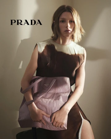 Model with blonde hair holding a light purple Prada tote bag wearing a brown dress against a wall with strong shadows