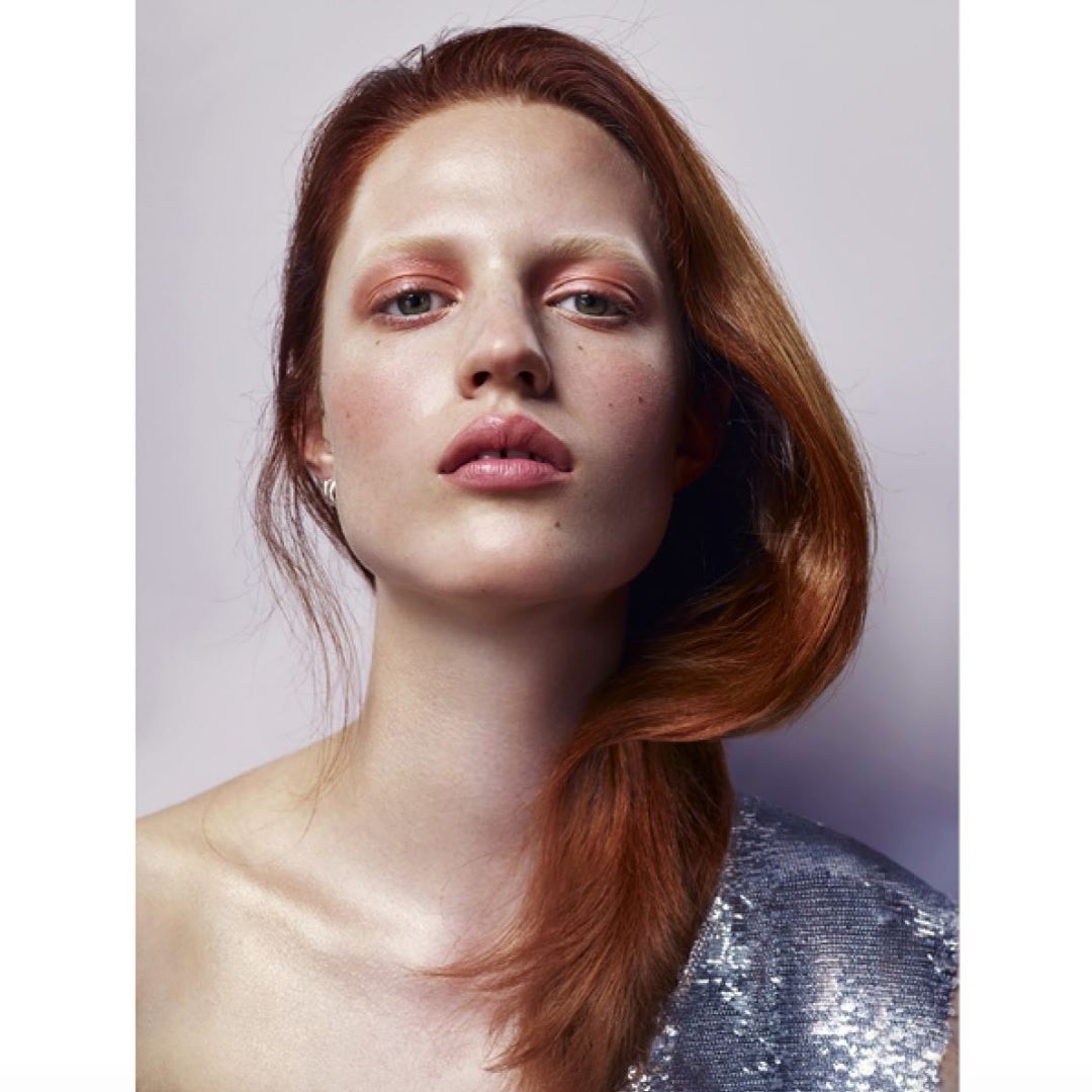 Close-up portrait of a fair-skinned adult woman with auburn hair, light pink makeup, and a silver sequin top against a soft purple background.