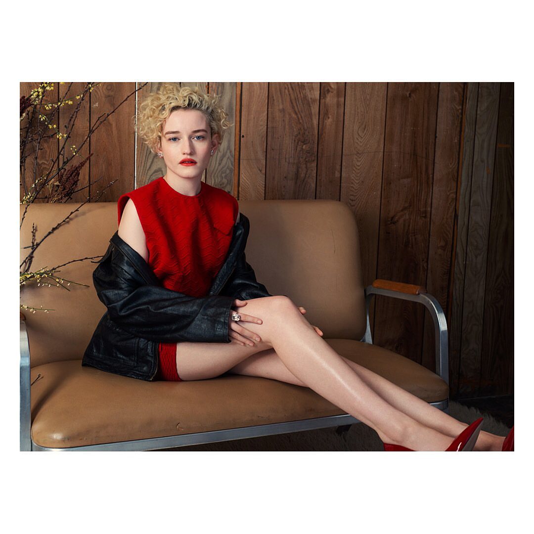 Julia Garner poses on a brown sofa in a red dress with a black jacket, and red heels, in an indoor studio setting with wood paneling.
