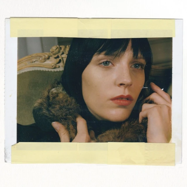 Close-up shot of a fair-skinned woman with short dark hair wearing a fur-trimmed black coat, seated on an antique chair in an indoor setting.