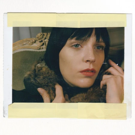 Close-up shot of a fair-skinned woman with short dark hair wearing a fur-trimmed black coat, seated on an antique chair in an indoor setting.