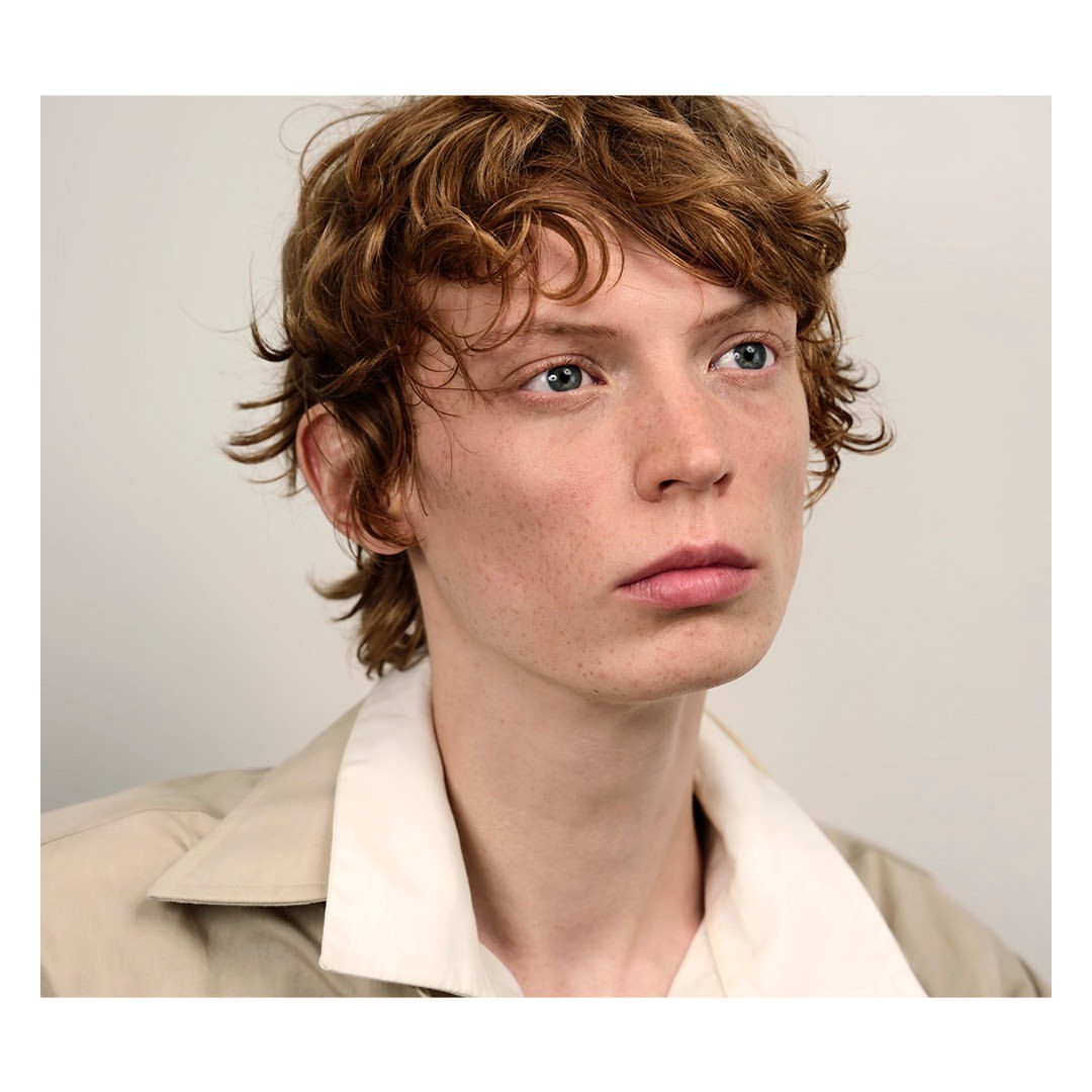 Close-up portrait of a young man with curly brown hair and blue eyes wearing a beige collared jacket against a plain light background