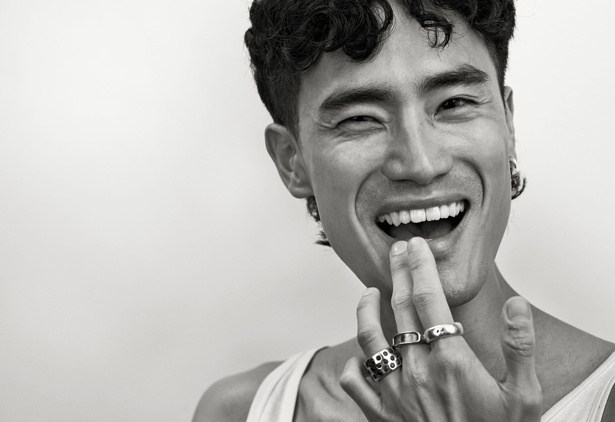 Close-up portrait of a smiling adult male of Asian descent with dark curly hair wearing jewelry against a plain background.