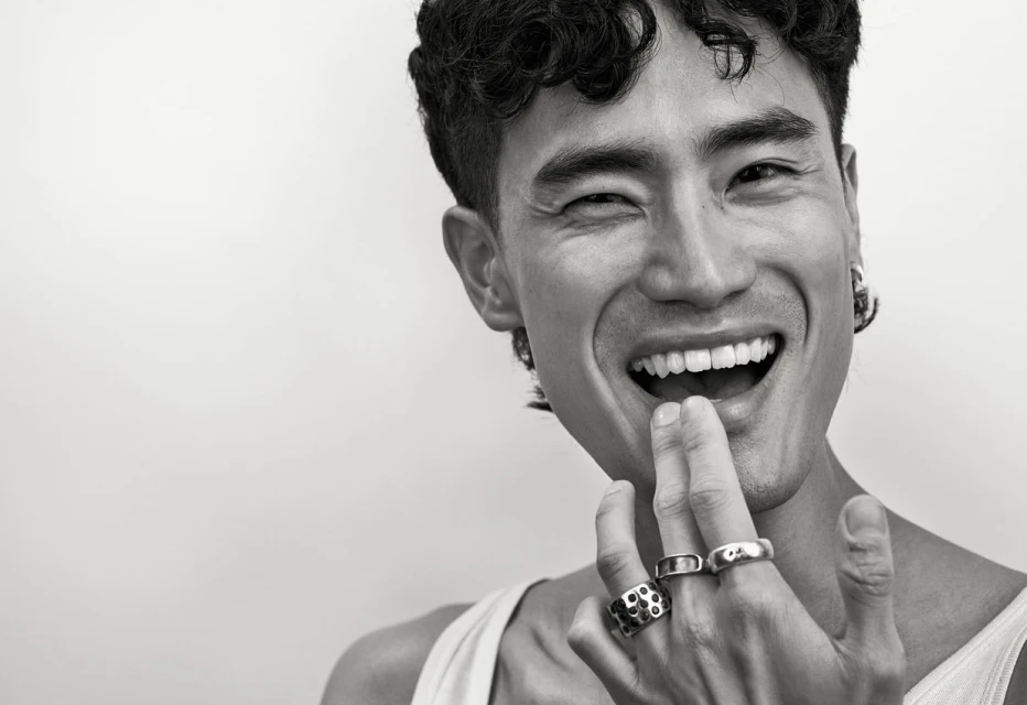 Close-up portrait of a smiling adult male of Asian descent with dark curly hair wearing jewelry against a plain background.