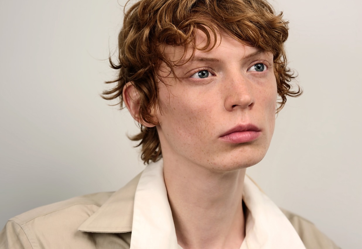 Headshot of a young adult male with fair skin, curly brown hair, and blue eyes wearing a tan collared shirt against a plain white background.