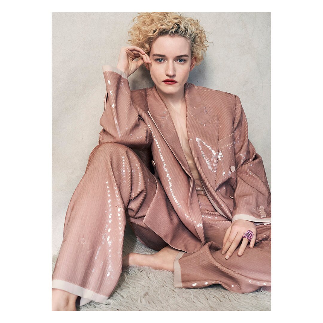 Julia Garner in a pink, sequined suit, seated on a white fur rug, gazing at the camera with a neutral expression.