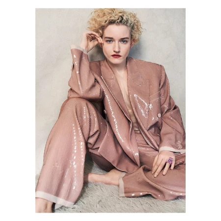 Julia Garner in a pink, sequined suit, seated on a white fur rug, gazing at the camera with a neutral expression.