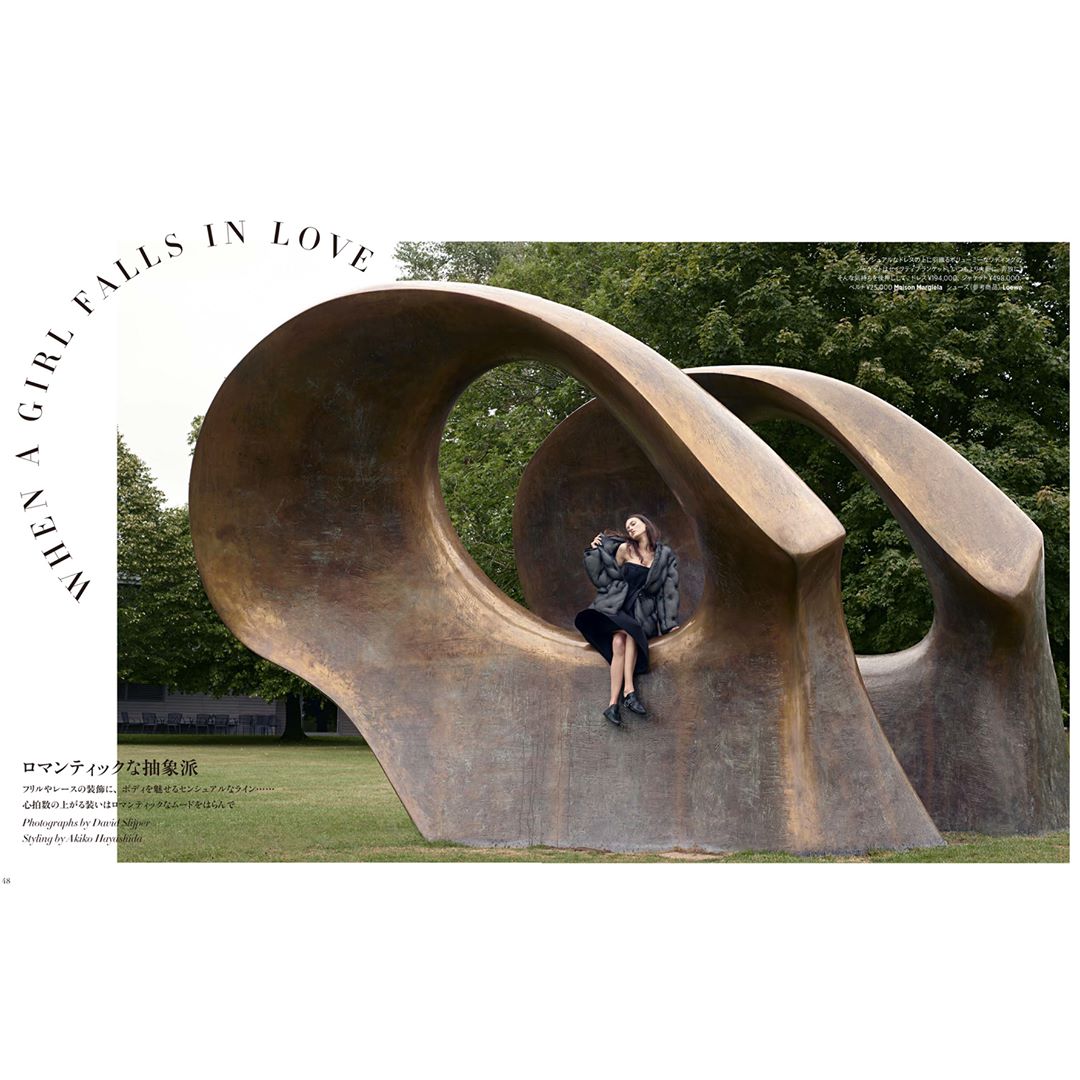 Woman sits on sculpture in black dress and jacket surrounded by green trees and grass on a sunny day with a book in hand.