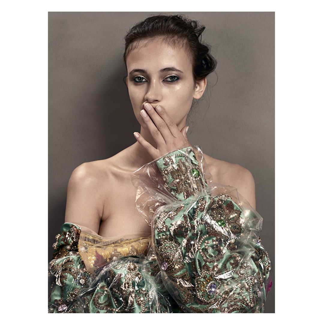 Striking portrait of a young woman with makeup and wrapped in layers of translucent plastic over ornate fabric, against a grey backdrop.