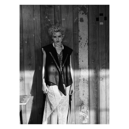 Model with curly blonde hair wearing a dark vest and patterned skirt against a weathered wood wall