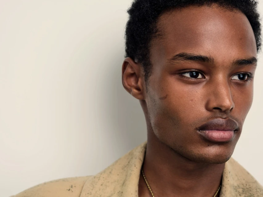 Close-up studio portrait of a young adult African-American man with short dark hair, wearing a beige coat and a gold chain.