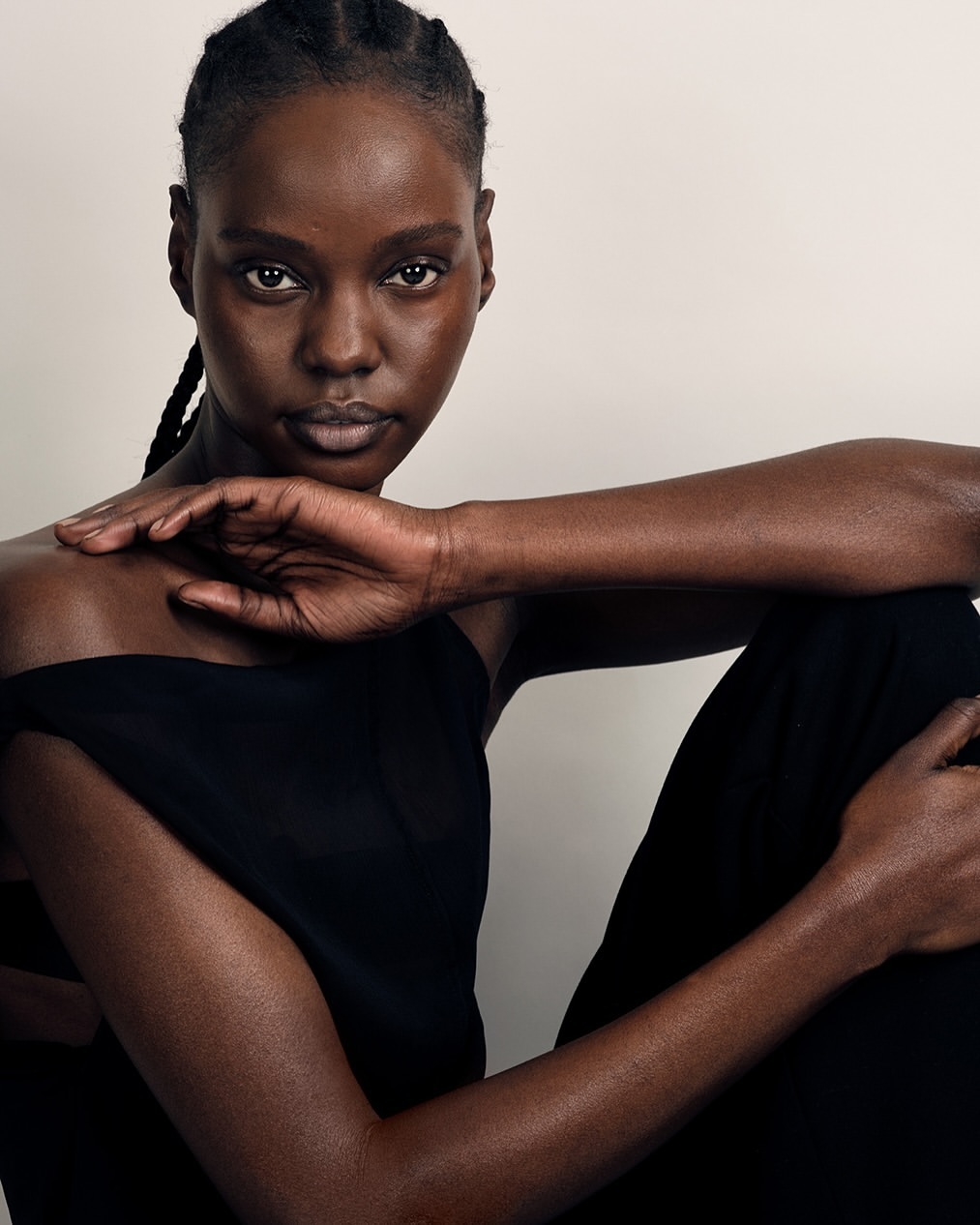 Portrait of a Black woman with braided hair, wearing a dark top, posing in a minimalist indoor studio setting with soft, diffused lighting.