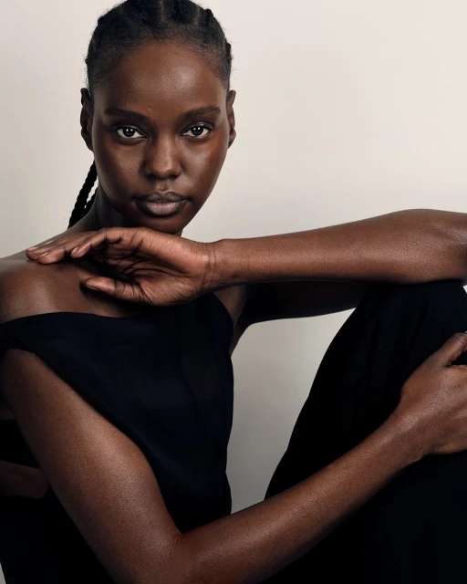 Portrait of a Black woman with braided hair, wearing a dark top, posing in a minimalist indoor studio setting with soft, diffused lighting.