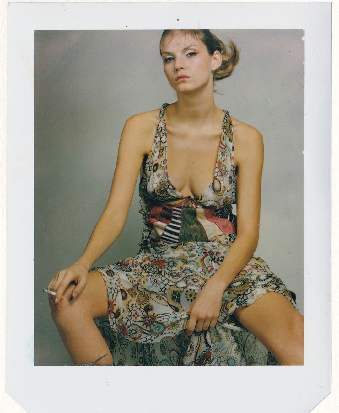 Young woman in floral dress sits in photo studio holding lit cigarette. Gray background and neutral tones create a classic portrait.