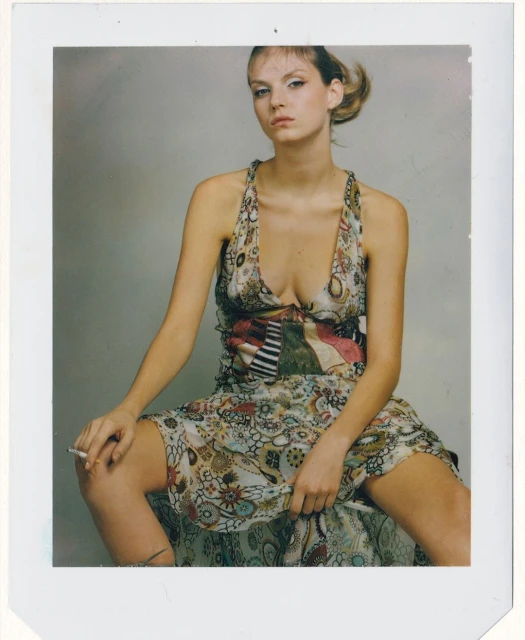 Young woman in floral dress sits in photo studio holding lit cigarette. Gray background and neutral tones create a classic portrait.