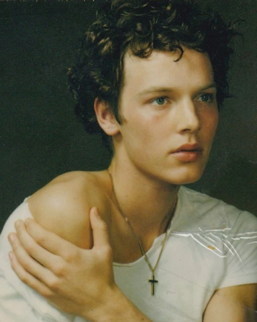 Close-up portrait of a young adult man with curly brown hair wearing a white shirt and a gold cross necklace against a dark background.