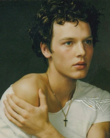 Close-up portrait of a young adult man with curly brown hair wearing a white shirt and a gold cross necklace against a dark background.