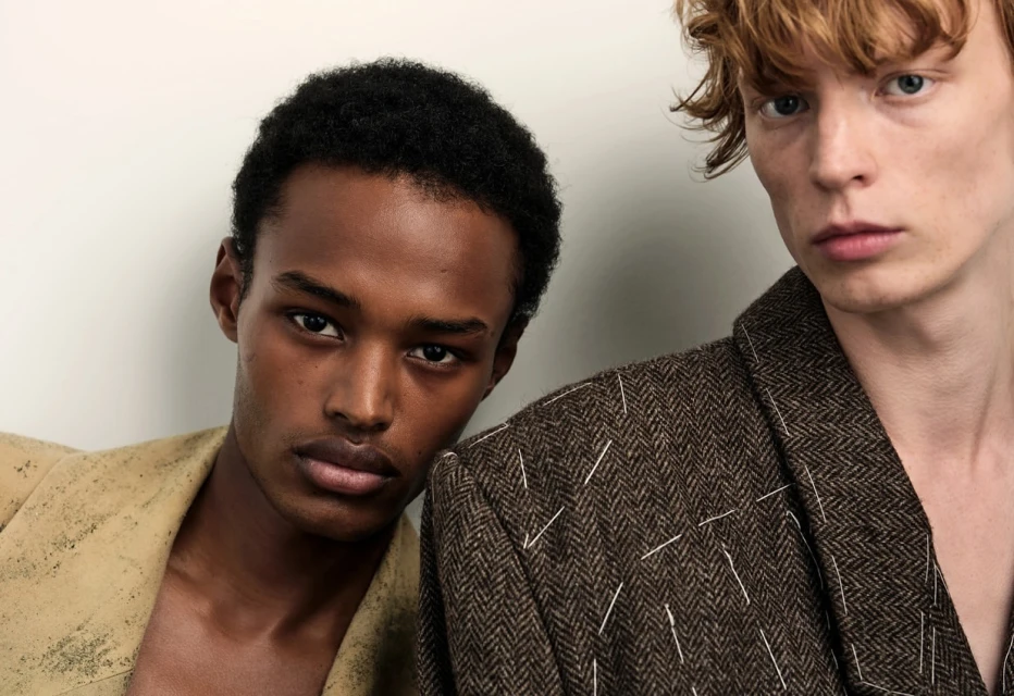 Two men in stylish blazers pose indoors against a white wall, showcasing fashion and portrait photography with soft lighting.