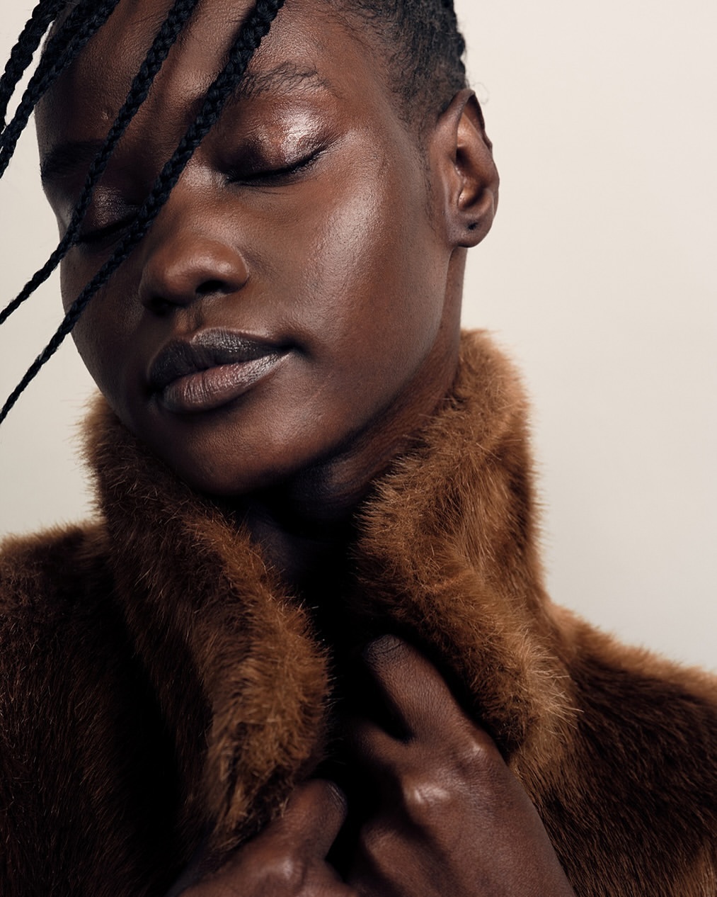 Close-up portrait of a Black woman with closed eyes wearing a brown textured faux fur coat against a plain light background.