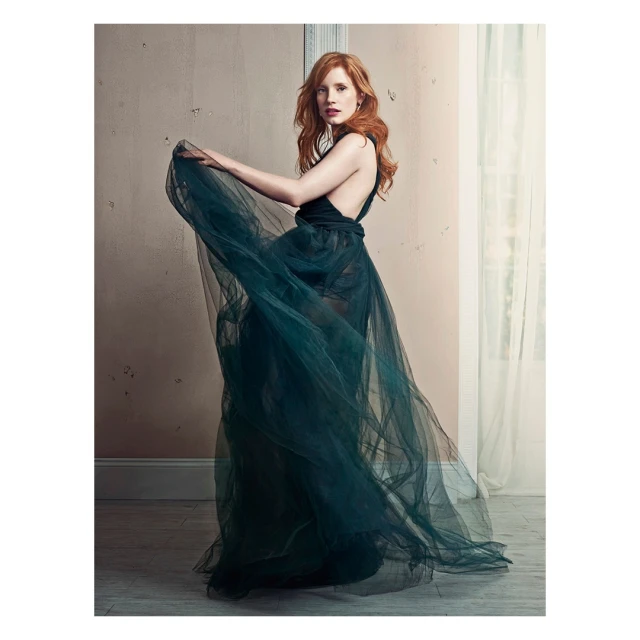 Jessica Chastain poses in a flowing green tulle gown in a photo studio with a neutral backdrop and natural light from a nearby window.