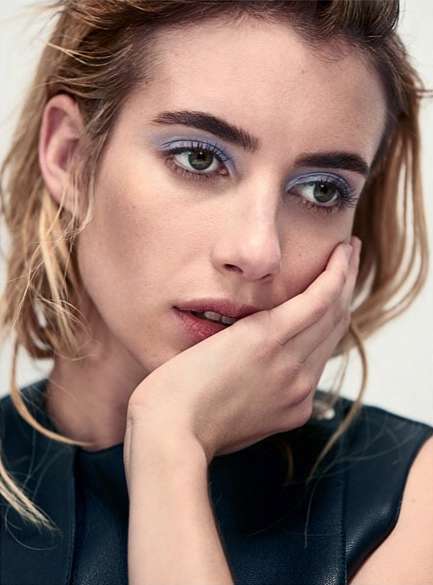Close-up of an adult female with blonde hair and green eyes wearing blue eye shadow and a dark blue top, resting her face on her hand.