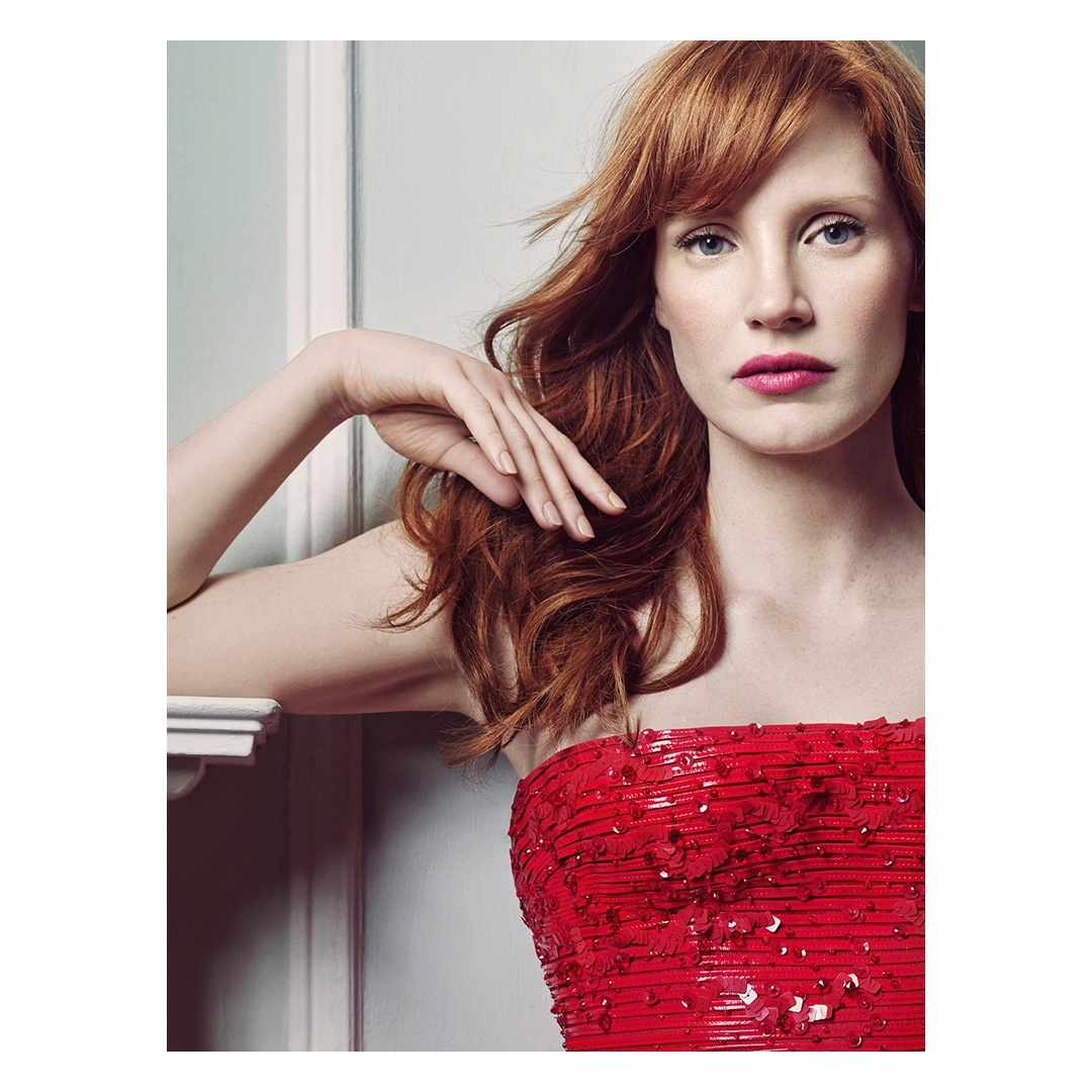 Close-up shot of an adult woman with long red hair, wearing a sequined strapless red dress, posing indoors in front of a white wall.