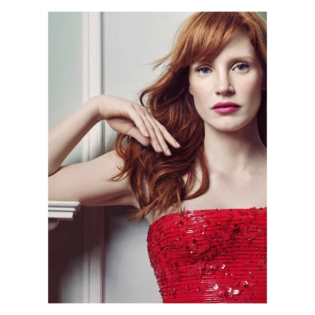 Close-up shot of an adult woman with long red hair, wearing a sequined strapless red dress, posing indoors in front of a white wall.