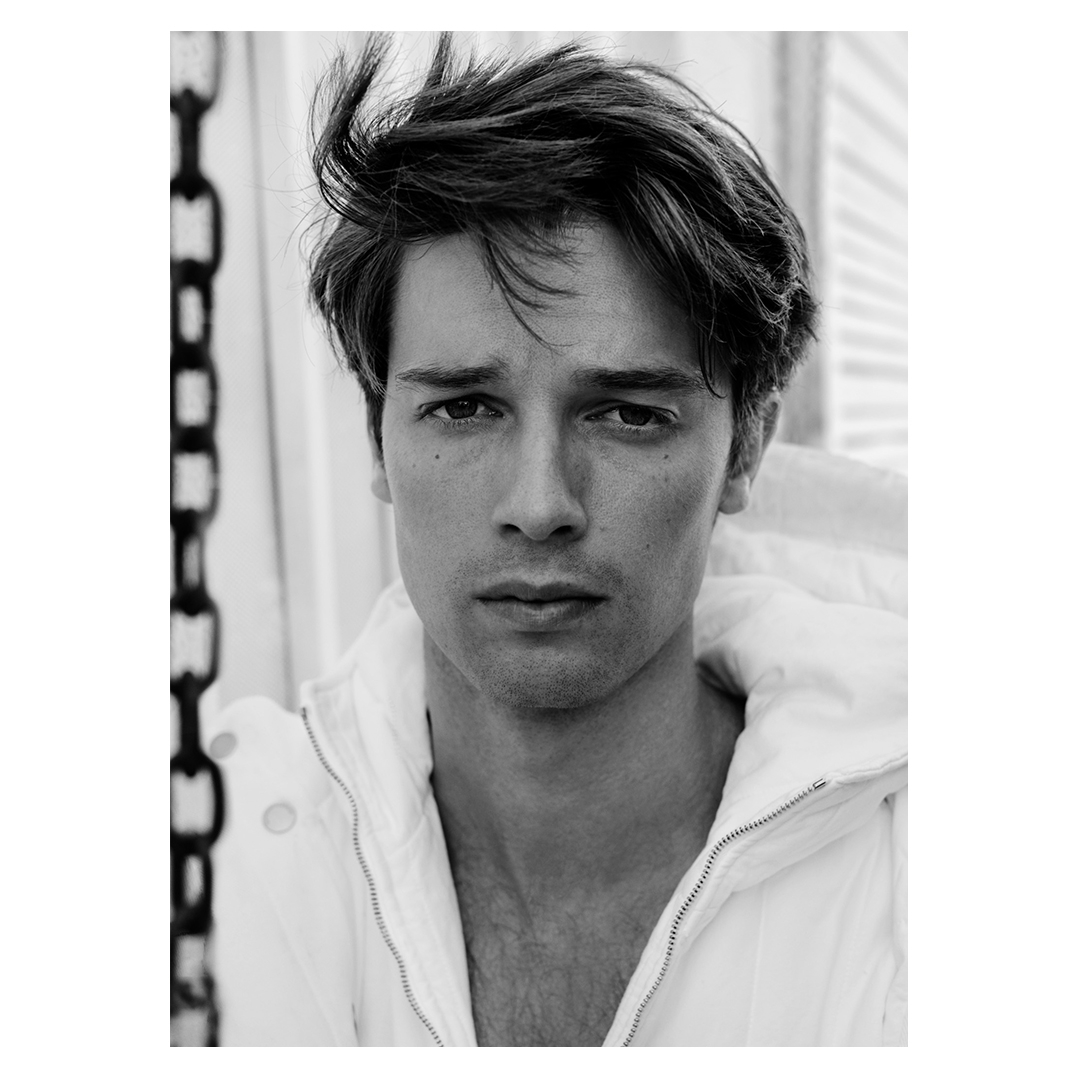 Portrait of a young adult male with dark hair, wearing a white hooded jacket in a monochromatic outdoor setting, with a serious facial expression.