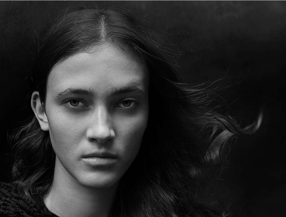 Portrait of a young adult woman with long, dark hair against a dark background, showcasing a close-up of her face and upper body.