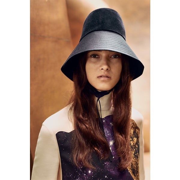 Portrait of a woman with long brown hair wearing a black hat and a white patterned shirt against a neutral-toned background.