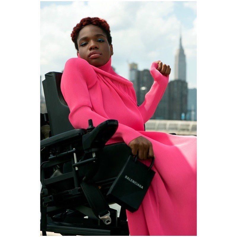 Portrait of female in pink gown seated in wheelchair, holding a black Balenciaga purse with city skyline in the background.