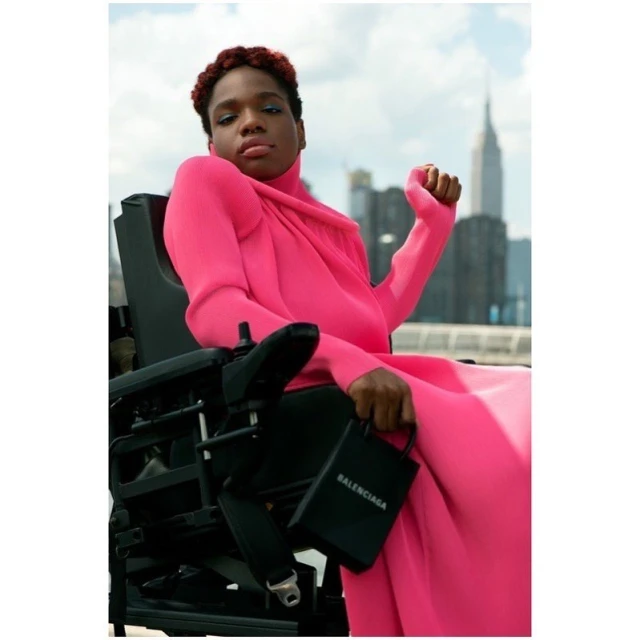 Portrait of female in pink gown seated in wheelchair, holding a black Balenciaga purse with city skyline in the background.