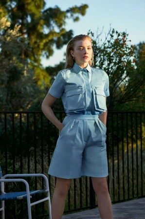 Adult woman poses outdoors wearing a blue short-sleeved button-up shirt and matching Bermuda shorts with hands in pockets.