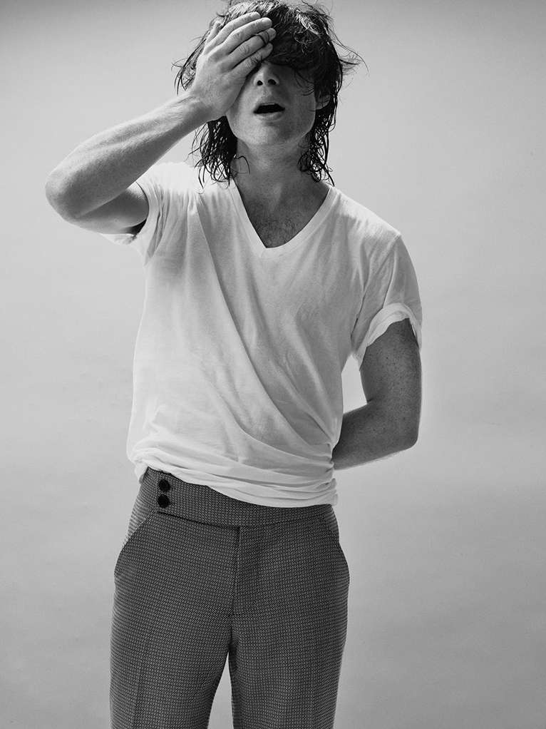 Monochrome portrait of a male model in a white t-shirt and patterned pants covering his face with his hand in a studio setting.